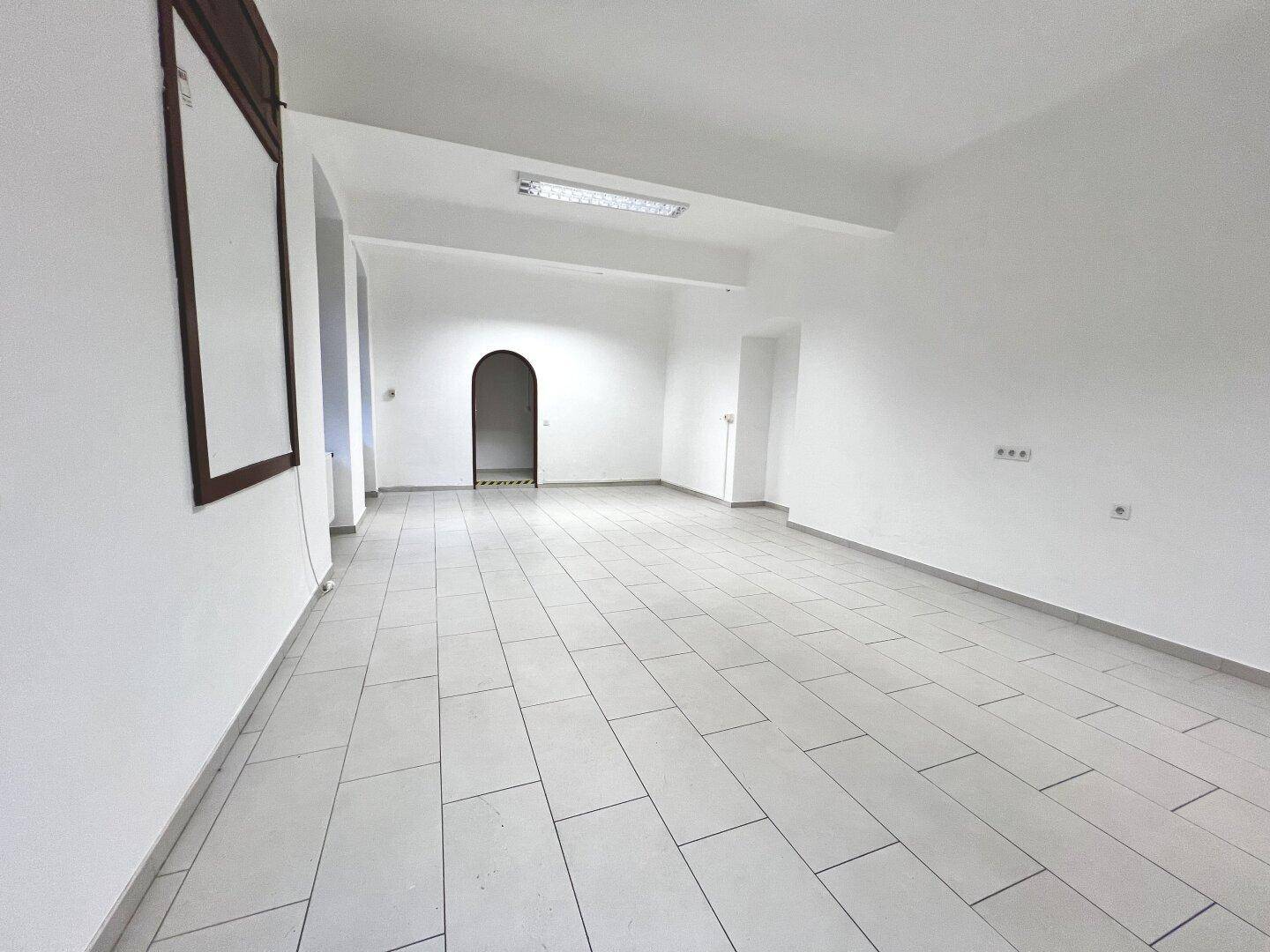 Großer, leerer Raum mit weißen Wänden, Fliesenboden und einem Durchgang mit Rundbogen.
