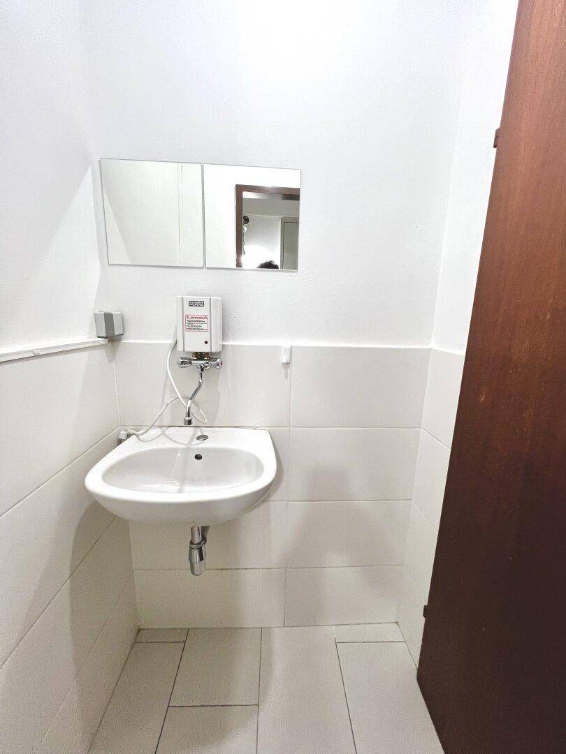 Kleiner Waschbereich mit weißem Waschbecken, Spiegel und hellen Fliesen an den Wänden.