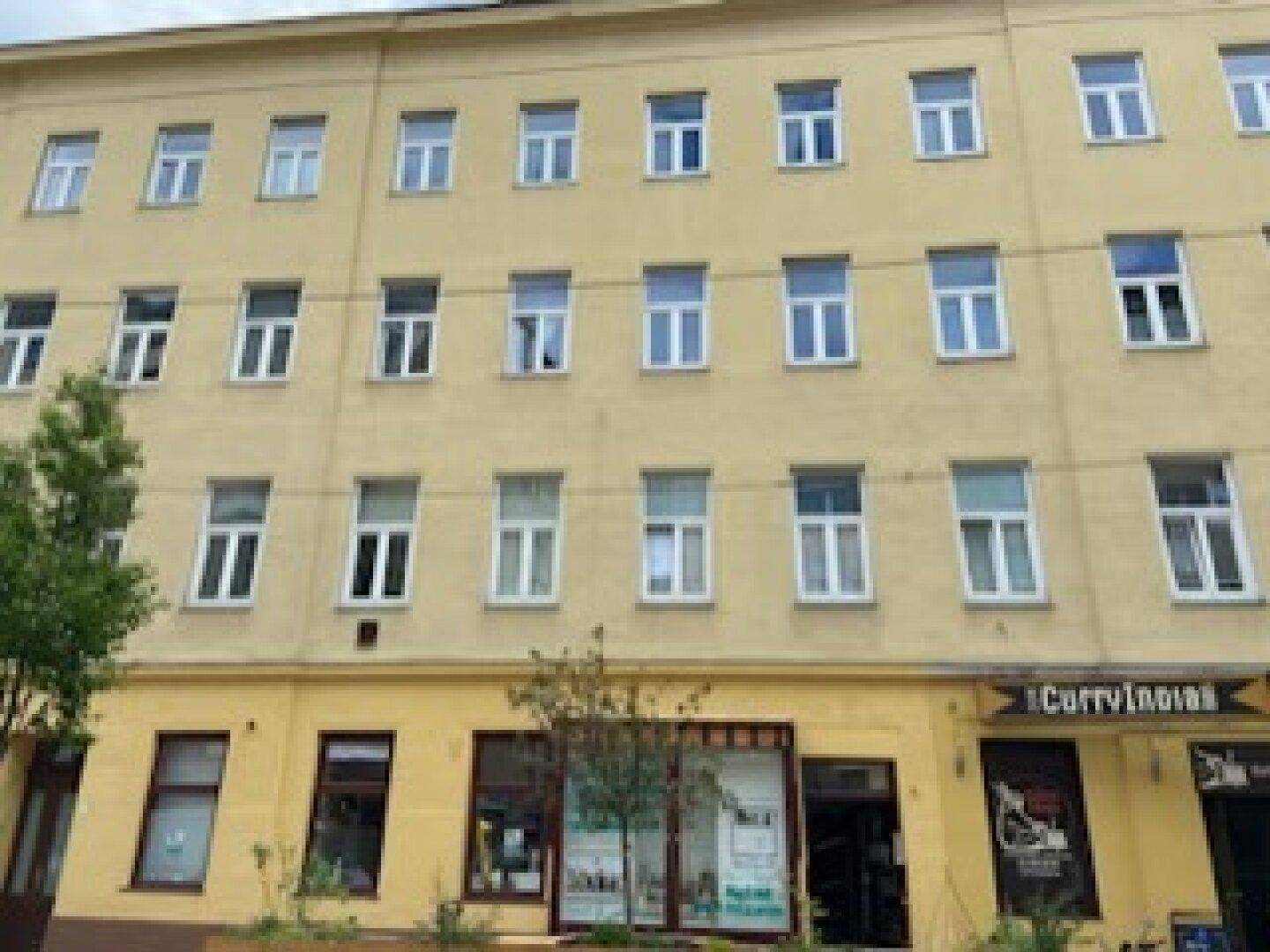 Fassade eines mehrstöckigen Altbaus mit vielen Fenstern und Erdgeschossgeschäften an einer Straße.