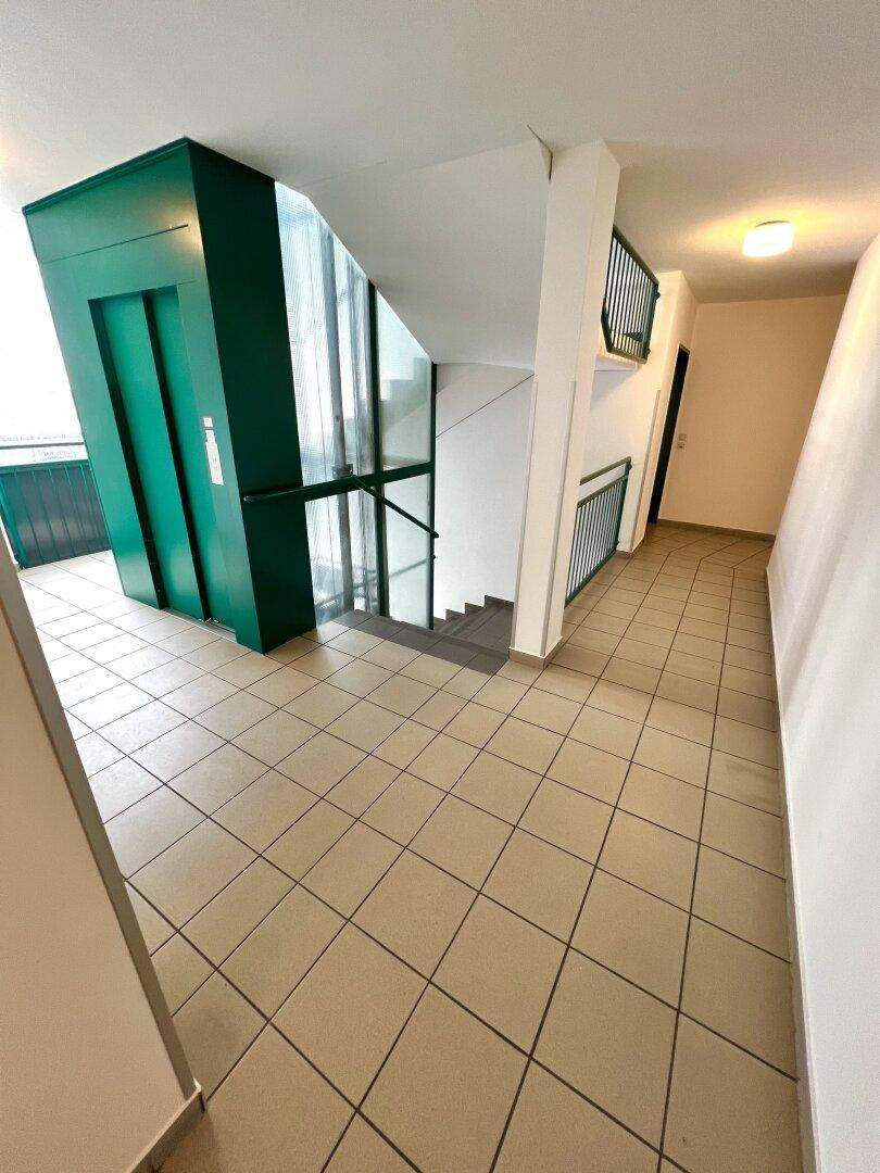 Heller Flur mit beigem Fliesenboden, einer Treppe und einem modernen grünen Aufzug.