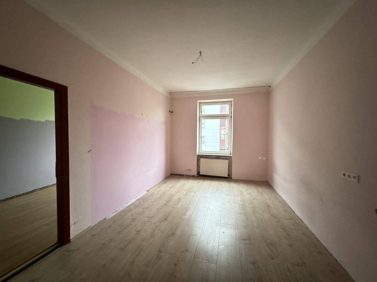 Leeres Zimmer mit rosa gestrichenen Wänden, Laminatboden und einem Fenster, das renovierungsbedürftig ist.