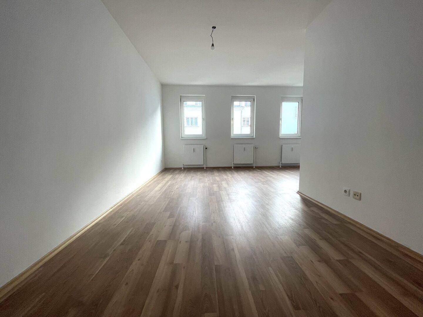 Helles, leeres Wohnzimmer mit Laminatboden, weißen Wänden und mehreren Fenstern.