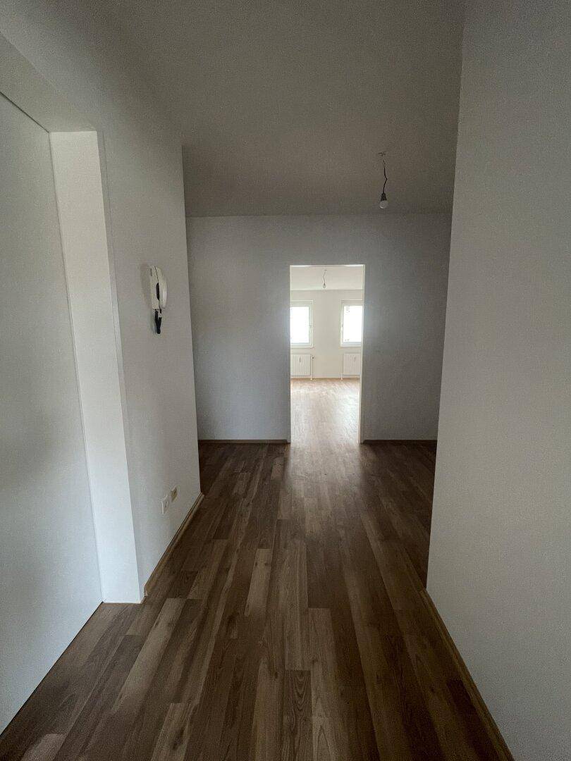 Langer Flur mit Laminatboden, weißen Wänden und Durchgang zu einem hellen Zimmer.