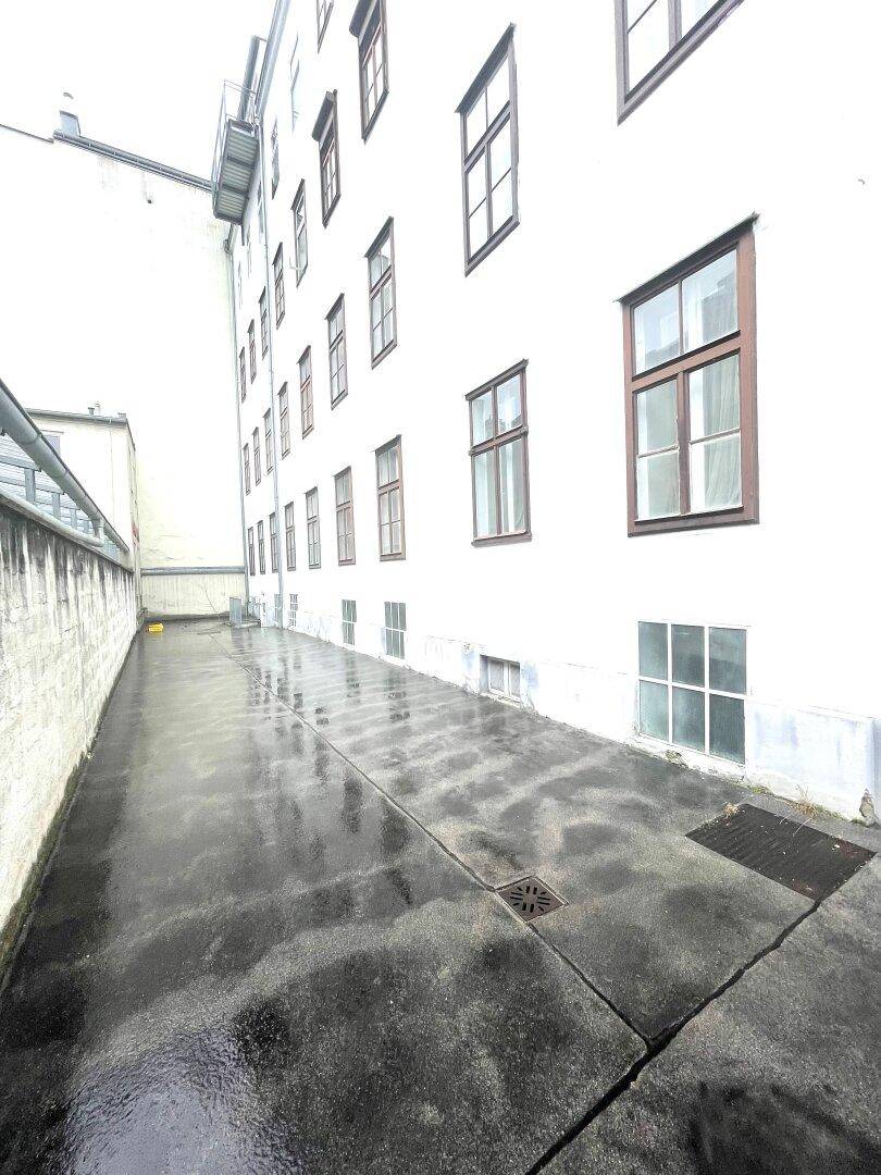Schmaler, nasser Betonweg entlang einer weißen Gebäudefassade mit vielen Fenstern.