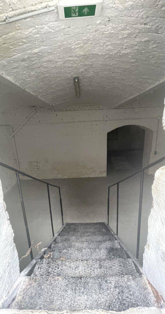 Blick von oben auf eine Betontreppe, die in einen dunkleren Bereich führt, mit Notausgangsschild.