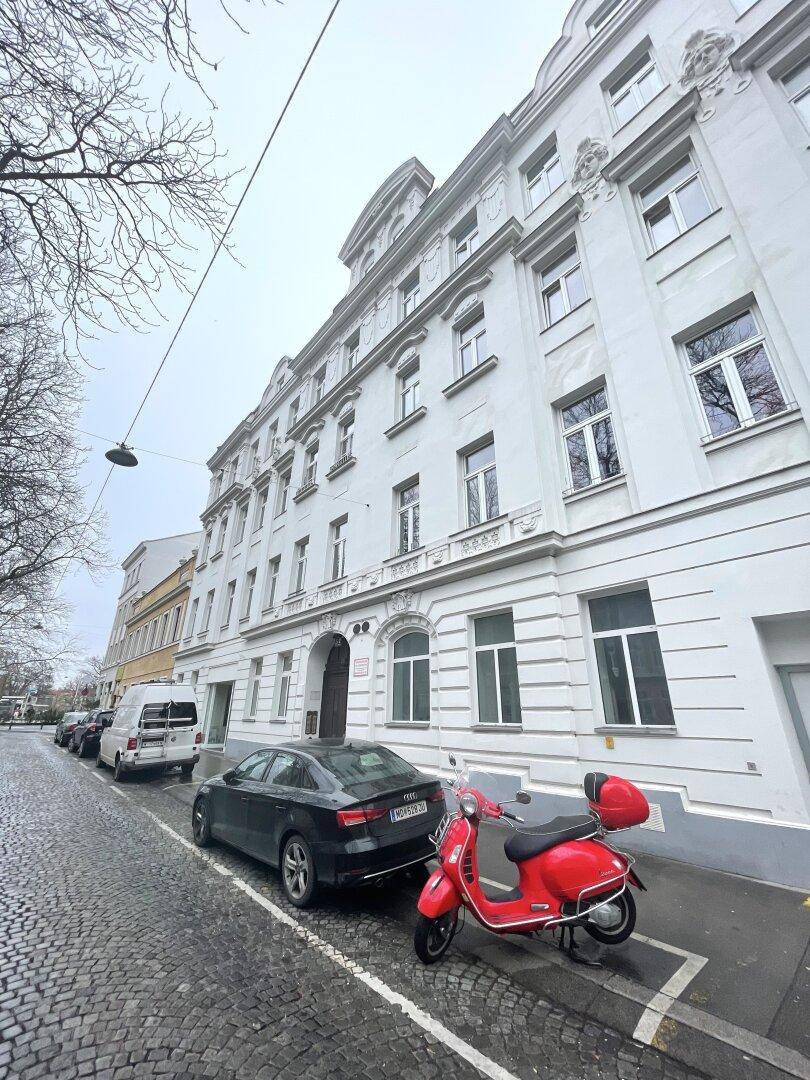 Historisches Gebäude mit klassischer Fassade und parkenden Autos auf der Kopfsteinpflasterstraße davor.