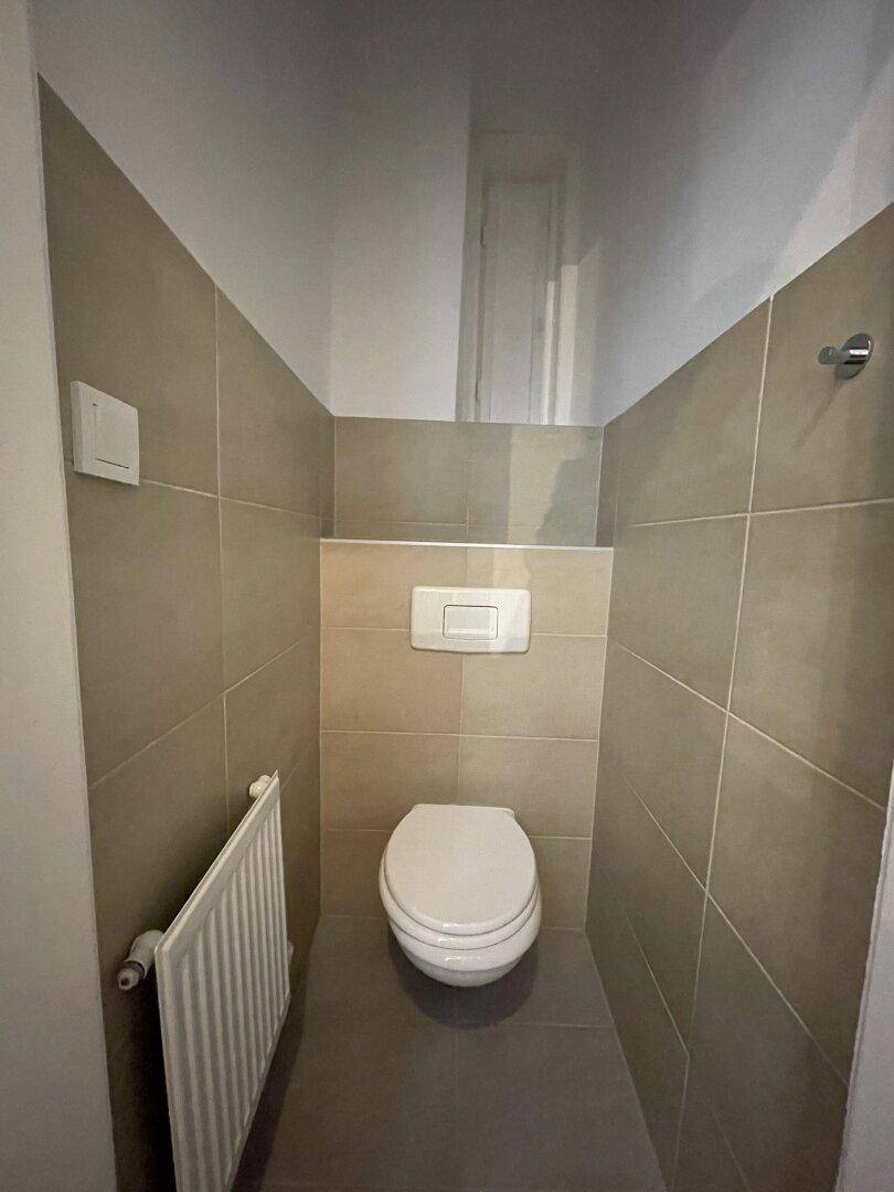 Separate Toilette mit beigefarbenen Fliesen, wandmontiertem WC und Heizkörper.