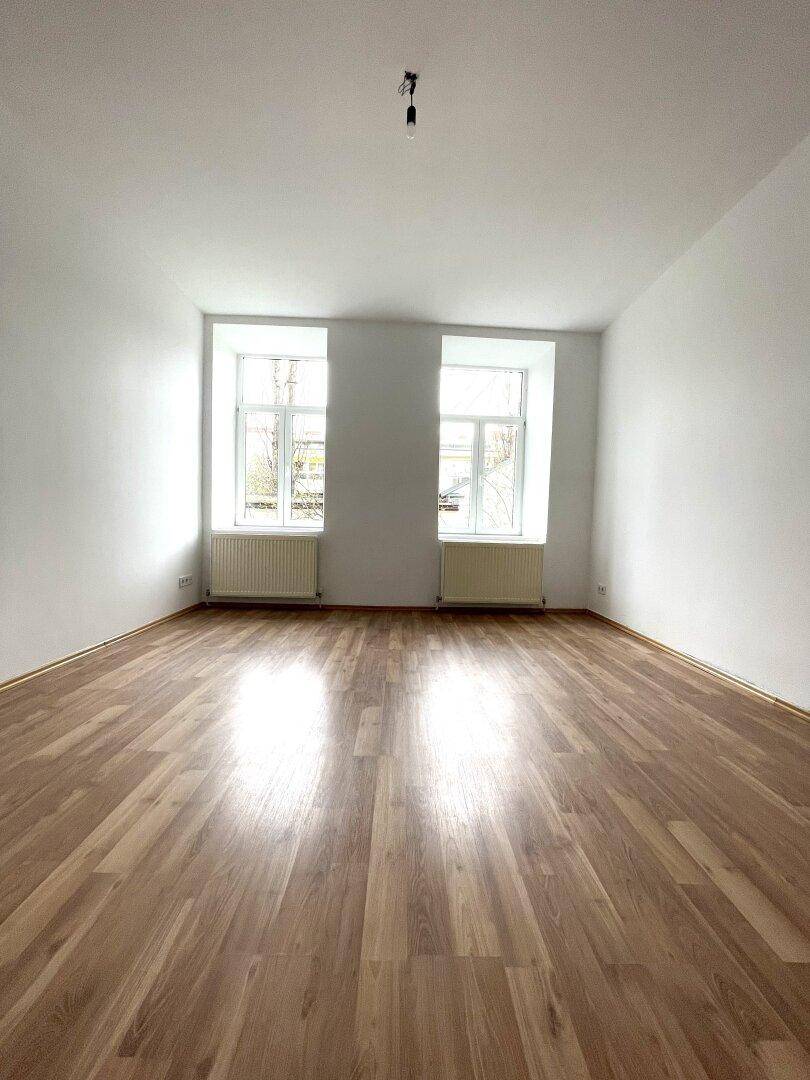 Heller Wohnraum mit Laminatboden, weißen Wänden und zwei großen Fenstern.