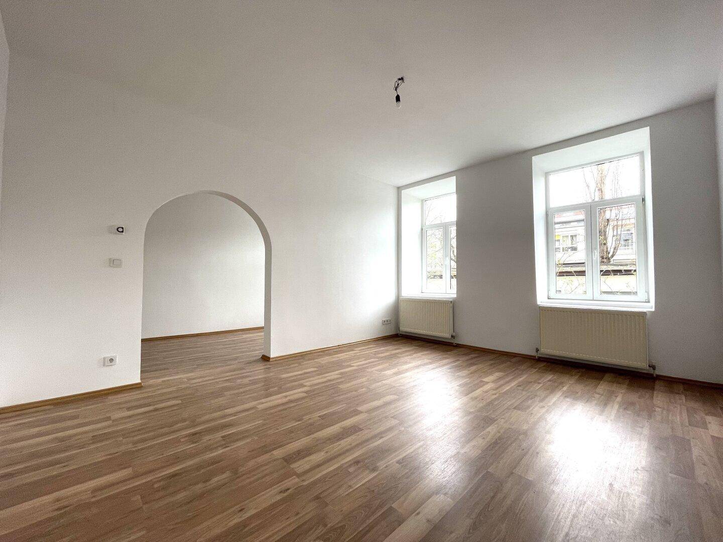 Geräumiger Wohnbereich mit Laminatboden, weiß gestrichenen Wänden und einem offenen Durchgang.