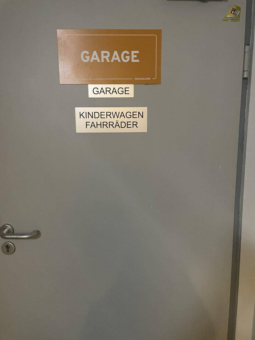Graue Tür mit Schildern, die auf eine Garage sowie einen Abstellraum für Kinderwagen und Fahrräder hinweisen.