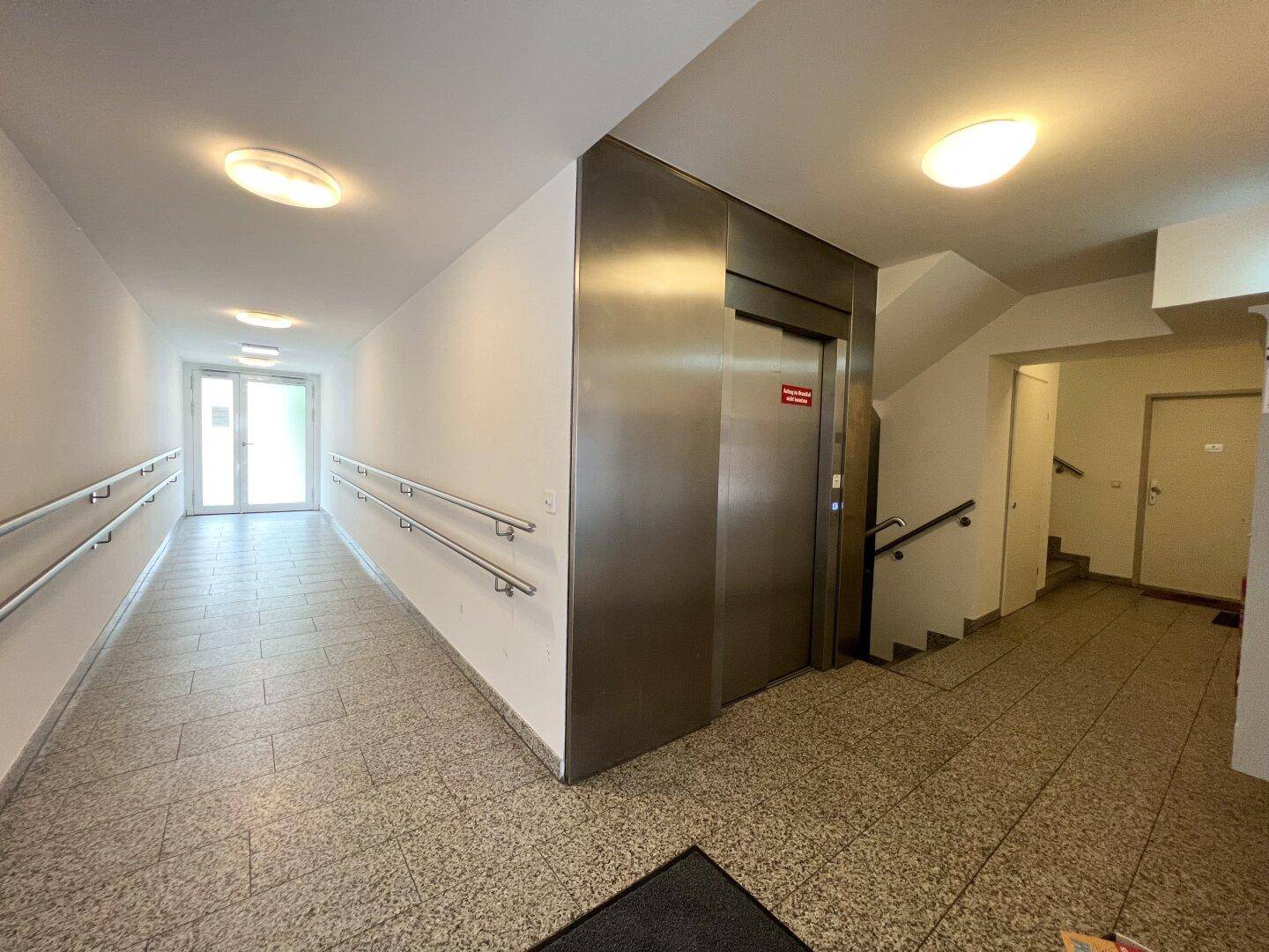 Geräumiger Flur im Gebäude mit Aufzug, Treppenhaus und Handläufen für Barrierefreiheit.