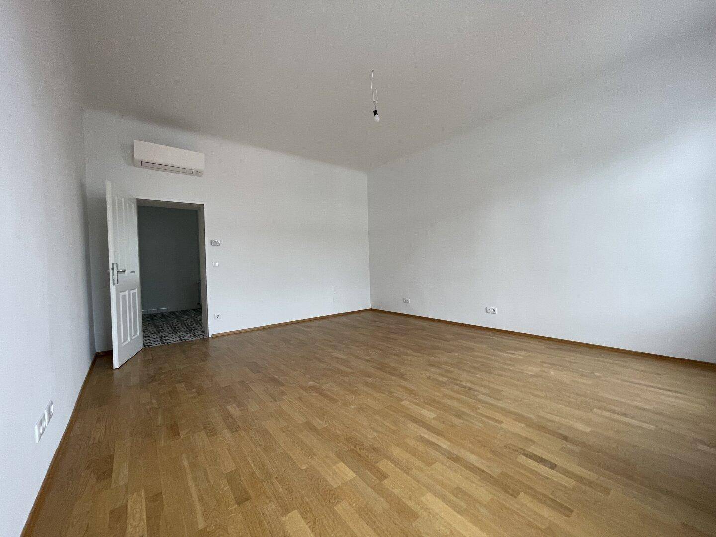Heller, leerer Raum mit Eichenparkettboden und Klimaanlage, der zu einem gefliesten Bereich führt.