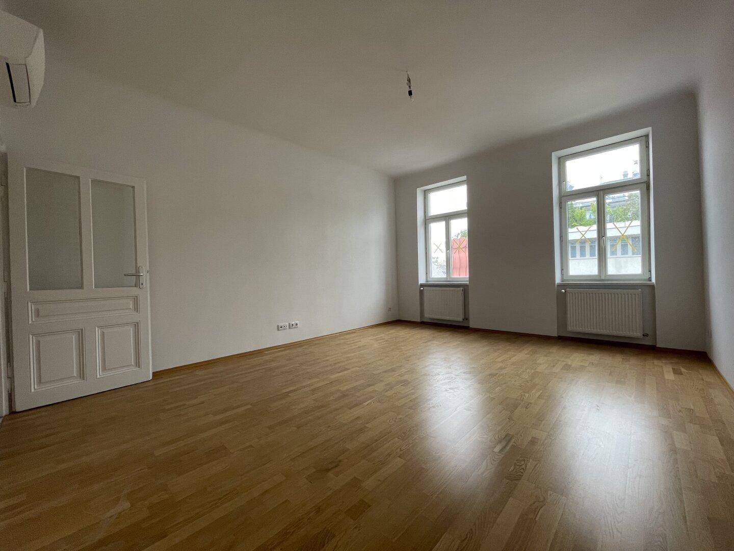 Geräumiger, heller Wohnbereich mit Holzboden, zwei Fenstern und Heizkörpern, bereit zur Einrichtung.