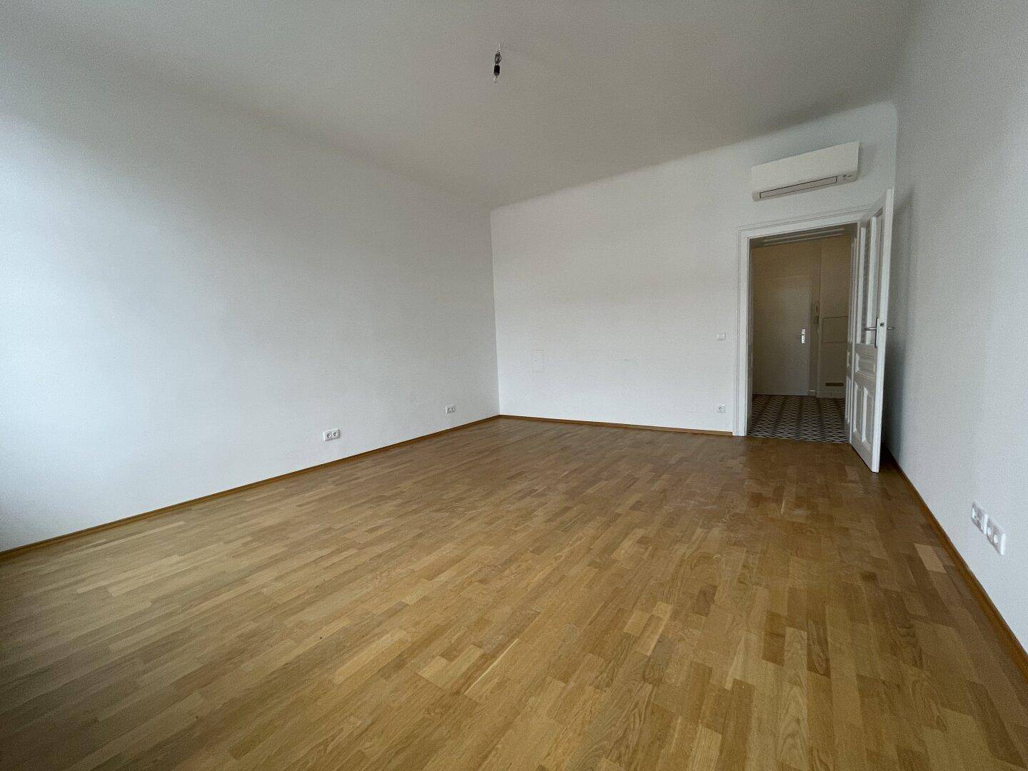 Leerer Wohnraum mit hellem Parkettboden und Klimaanlage, der einen Übergang zu einem gefliesten Bereich bietet.