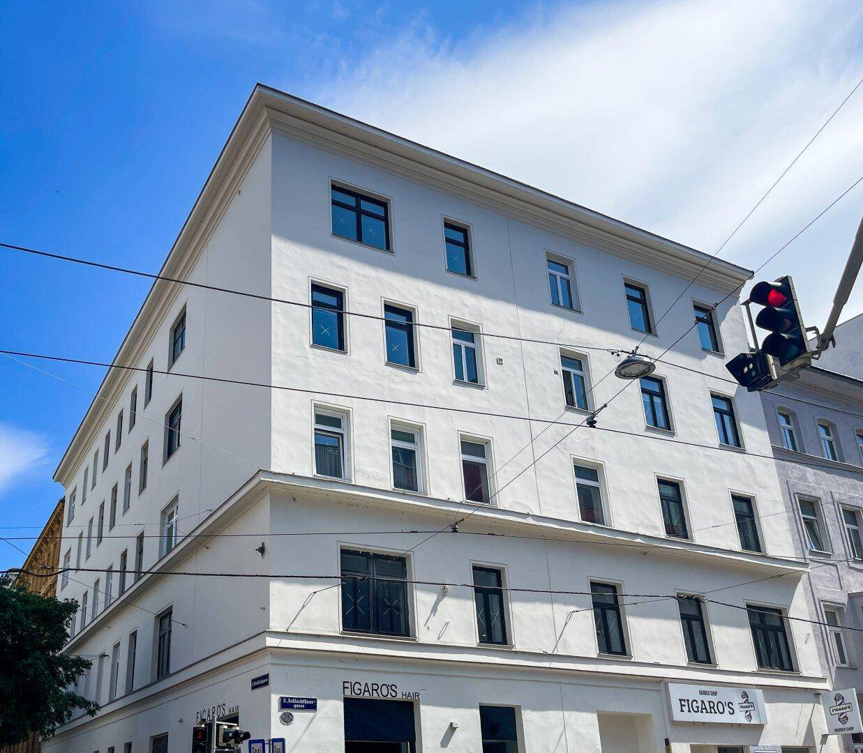 Weißes Mehrfamilienhaus mit vielen Fenstern und Geschäften im Erdgeschoss unter blauem Himmel.