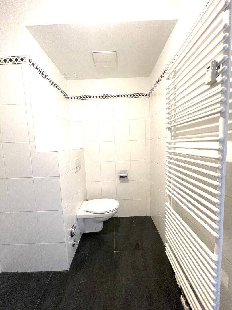 Separates WC mit wandhängender Toilette, beheiztem Handtuchhalter und dunklem Fliesenboden.