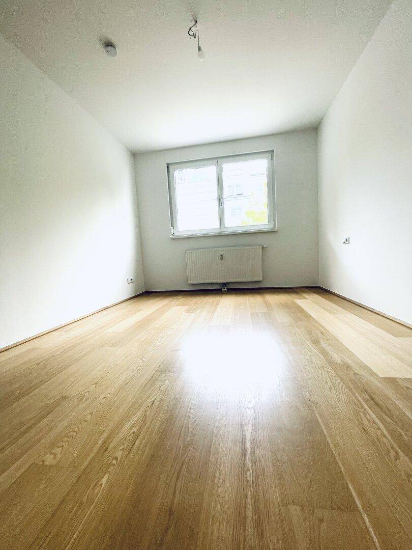 Helles, leeres Zimmer mit Parkettboden, weißen Wänden und einem Fenster mit Heizkörper.