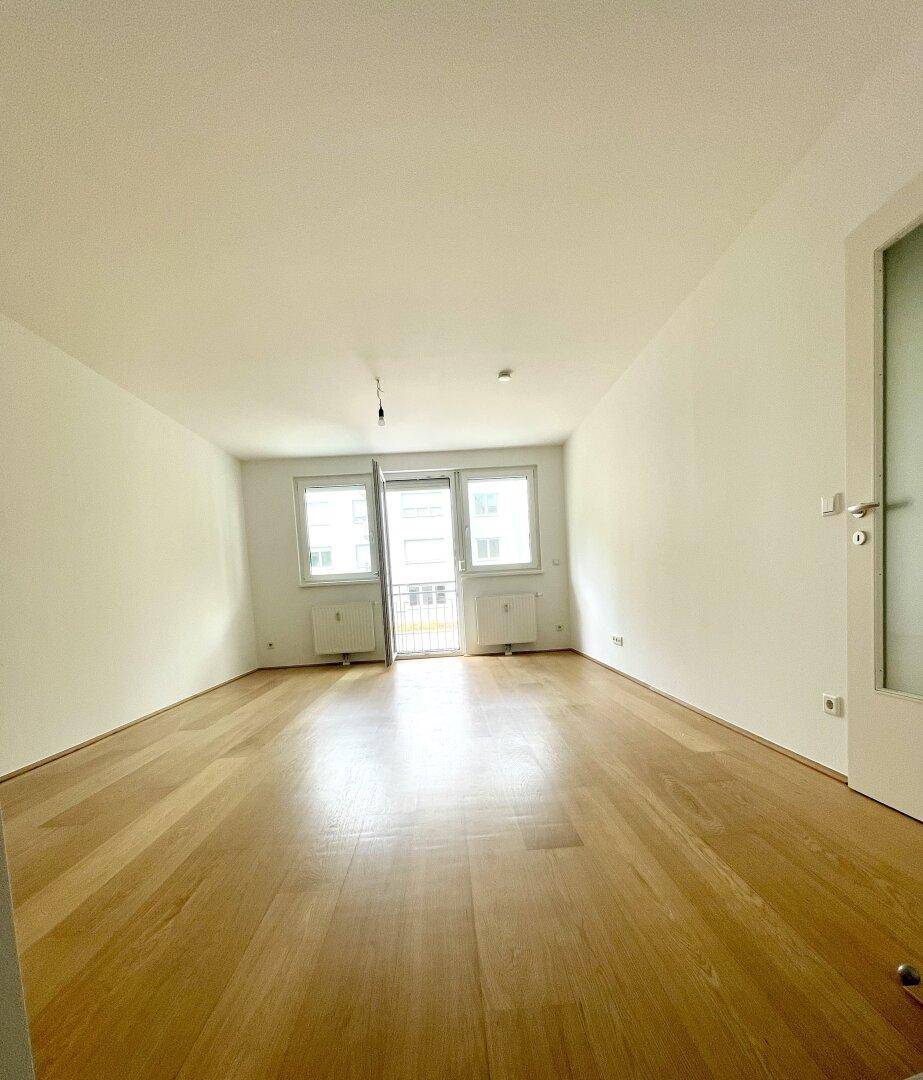Helles, leeres Wohnzimmer mit hellem Parkettboden und Zugang zu einem Balkon oder einer Terrasse.