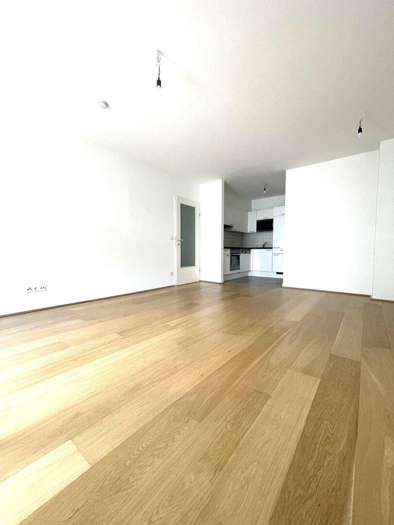 Geräumiges Wohnzimmer mit hellem Parkettboden und offenem Blick in die moderne Küche.