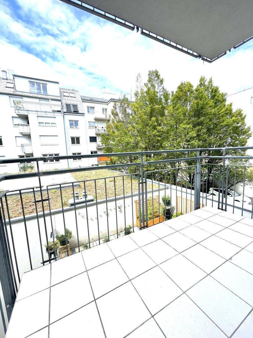 Heller Balkon mit Fliesenboden und Metallgeländer, bietet einen Blick auf Bäume und umliegende Gebäude.