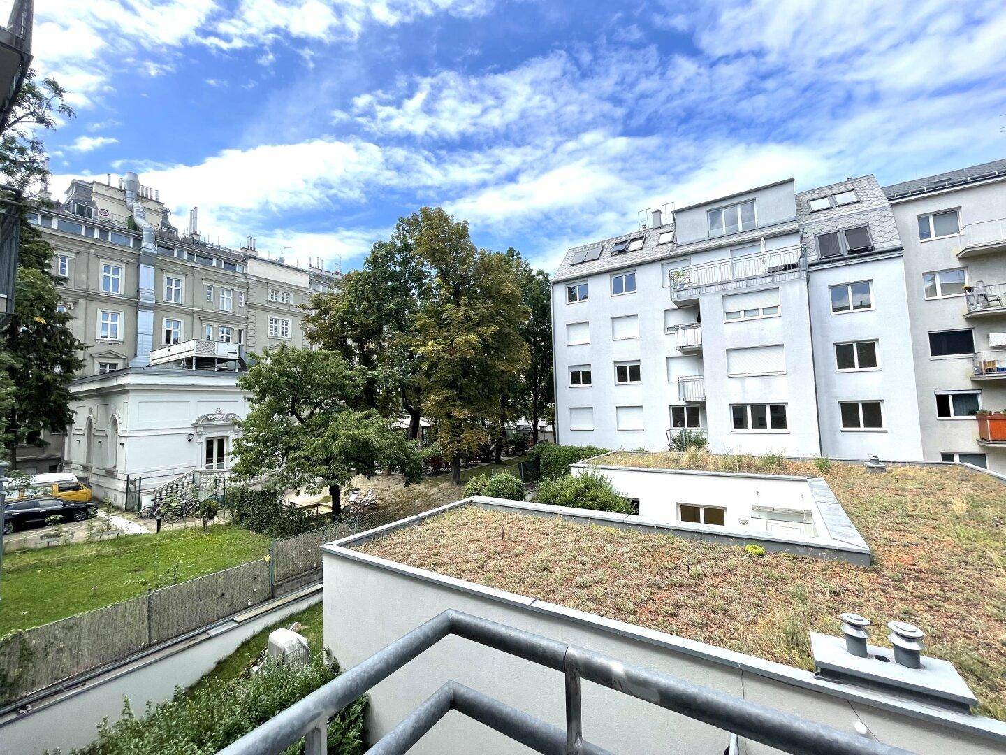 Ausblick vom Balkon auf ein begrüntes Dach, alte Bäume und weitere Wohngebäude in der Umgebung.