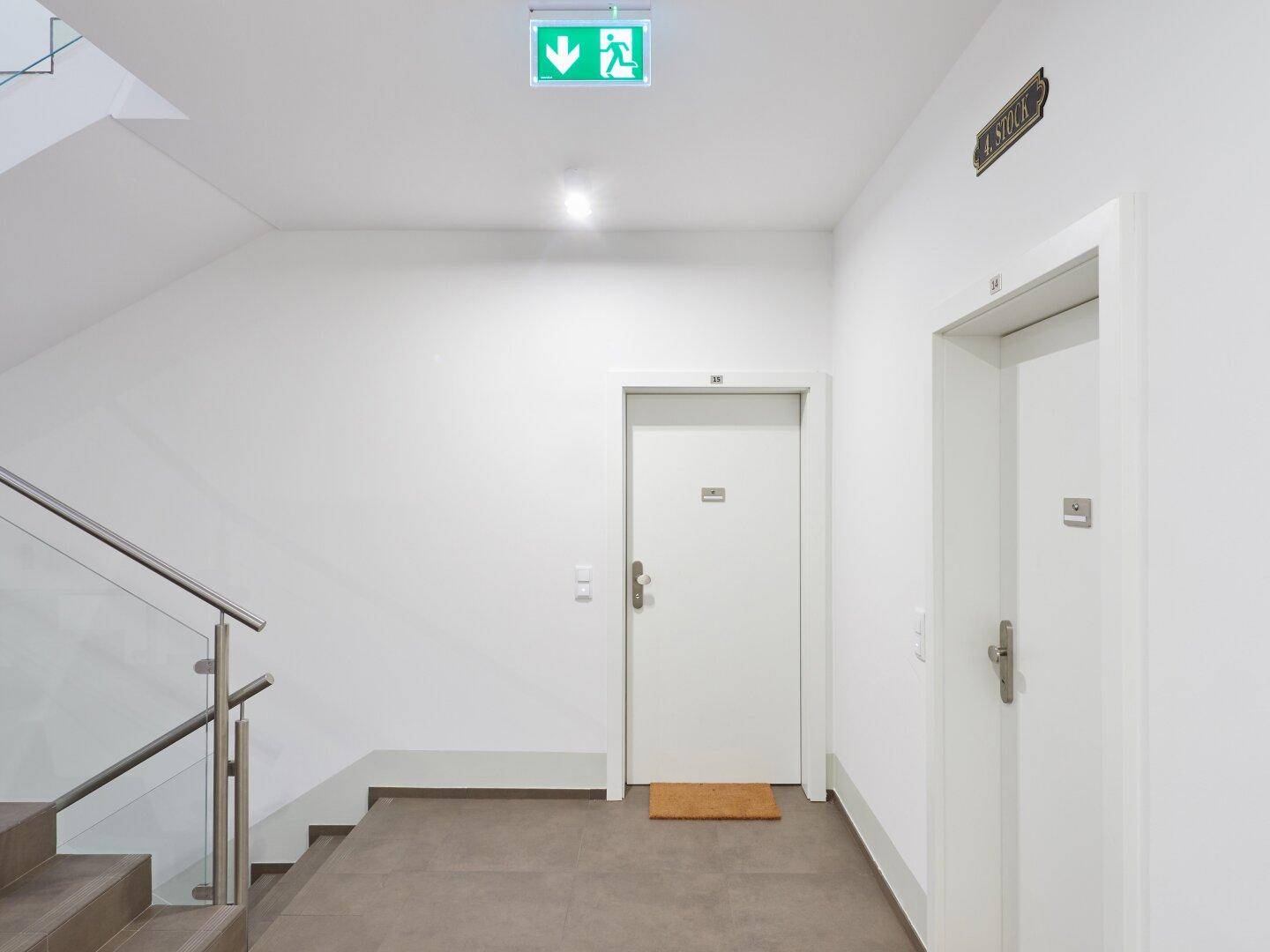 Moderner Treppenhausflur mit Notausgangsschild, Glastreppengeländer und Zugang zu den Wohneinheiten.
