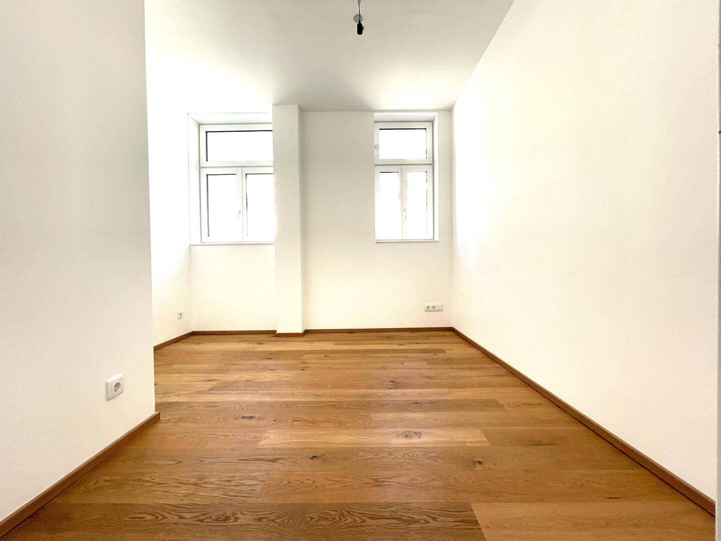 Helles, leeres Zimmer mit Eichenparkettboden und zwei großen Fenstern, ideal als Schlafzimmer.