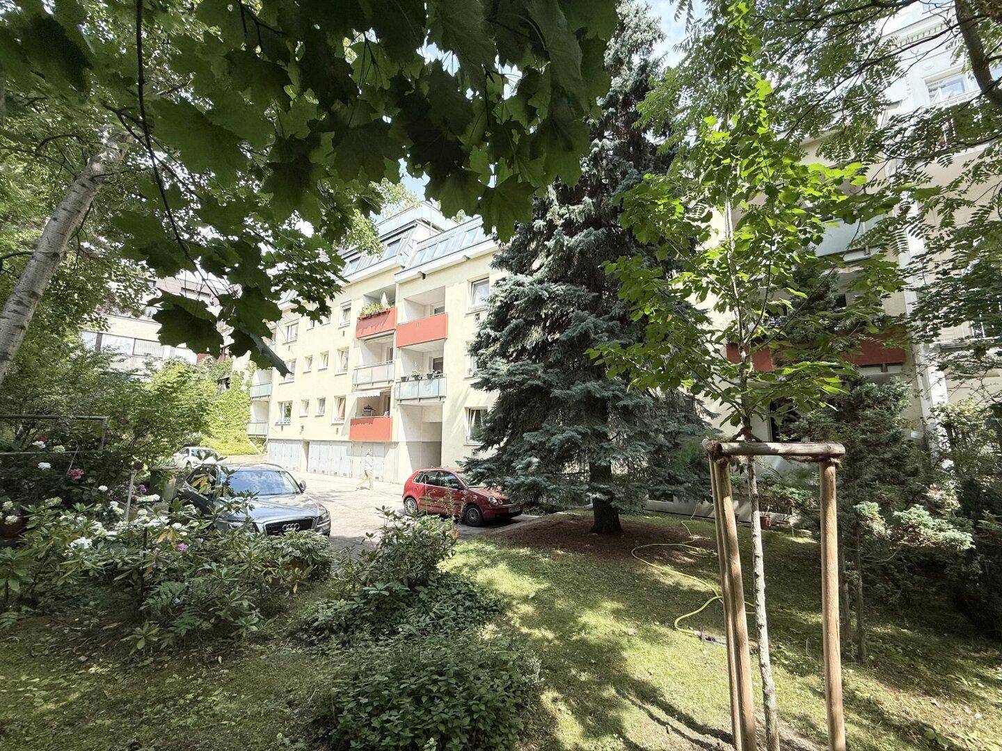 Mehrfamilienhaus mit Balkonen, umgeben von viel Grün und Bäumen sowie geparkten Autos.