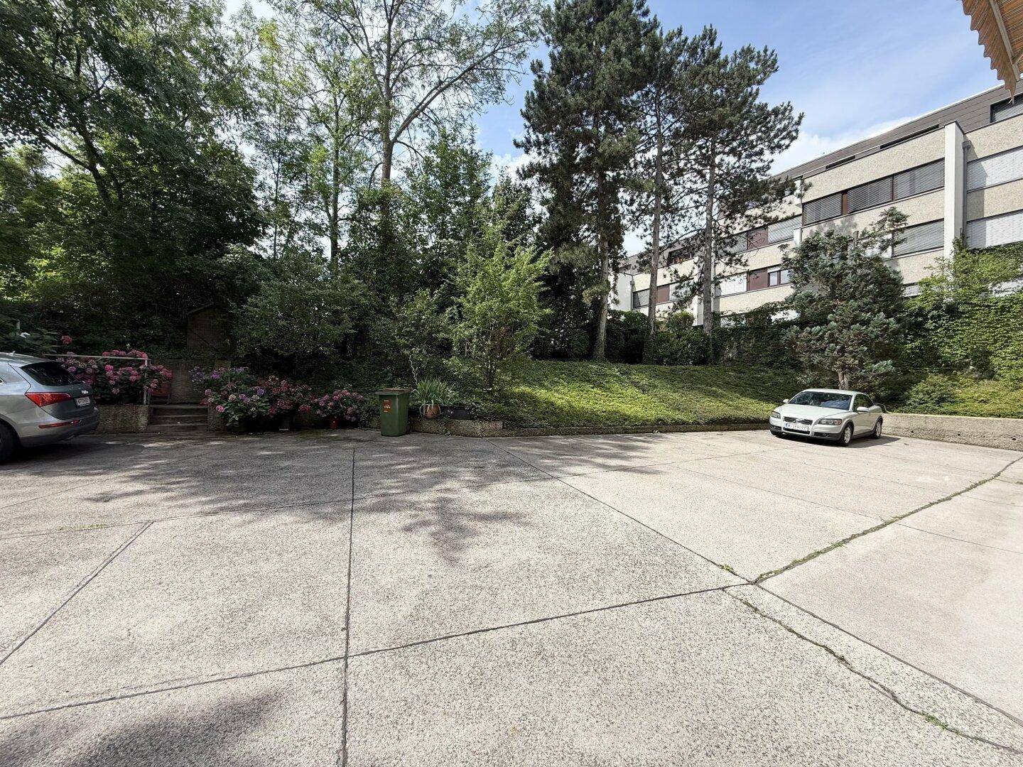 Außenparkplatz mit Betonfläche, umgeben von Bäumen und blühenden Sträuchern.
