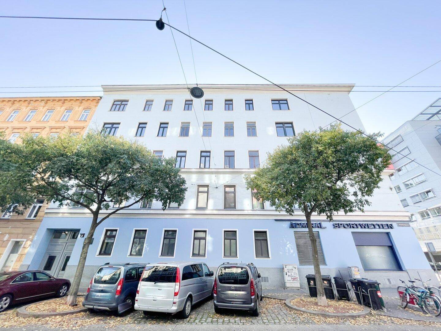 Gebäudefassade mit hellblauer und weißer Farbgebung, umgeben von Bäumen und parkenden Autos auf der Straße.