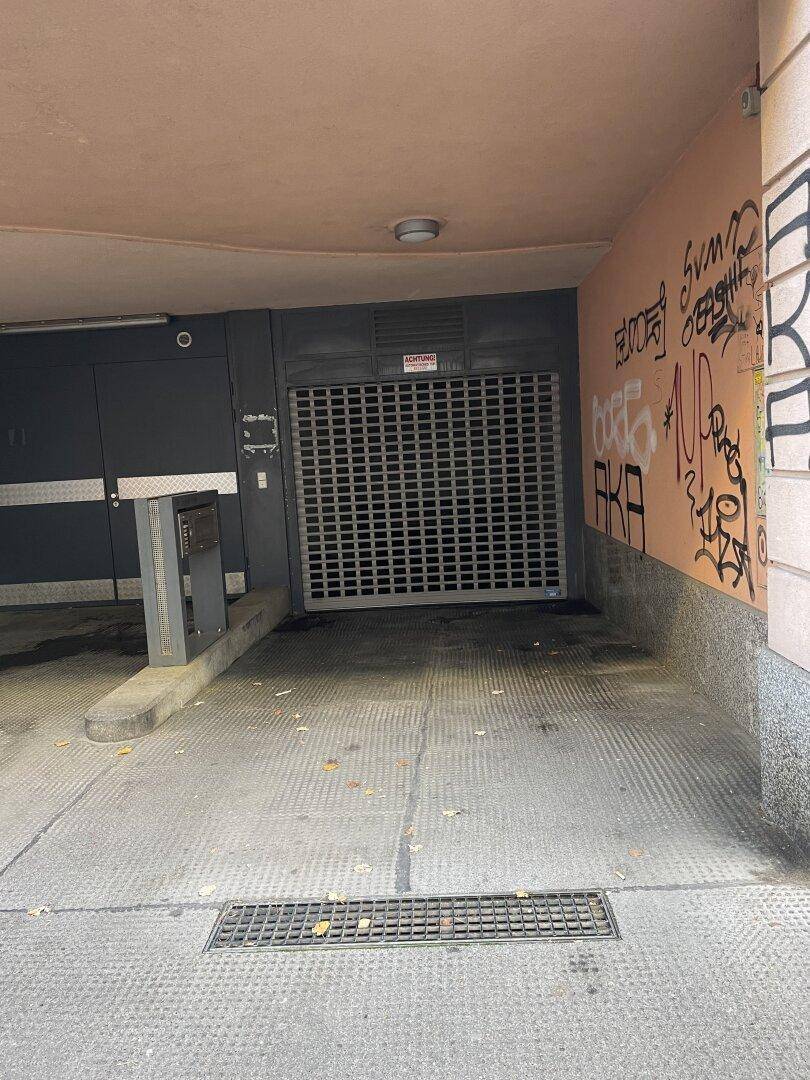 Einfahrt zur Tiefgarage mit geschlossenem Rolltor und Graffiti an der Wand, von außen gesehen.