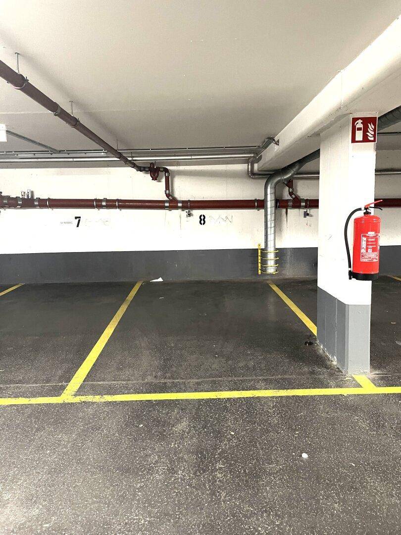 Leere Stellplätze in einer Tiefgarage mit sichtbaren Rohren an der Decke und einem Feuerlöscher.