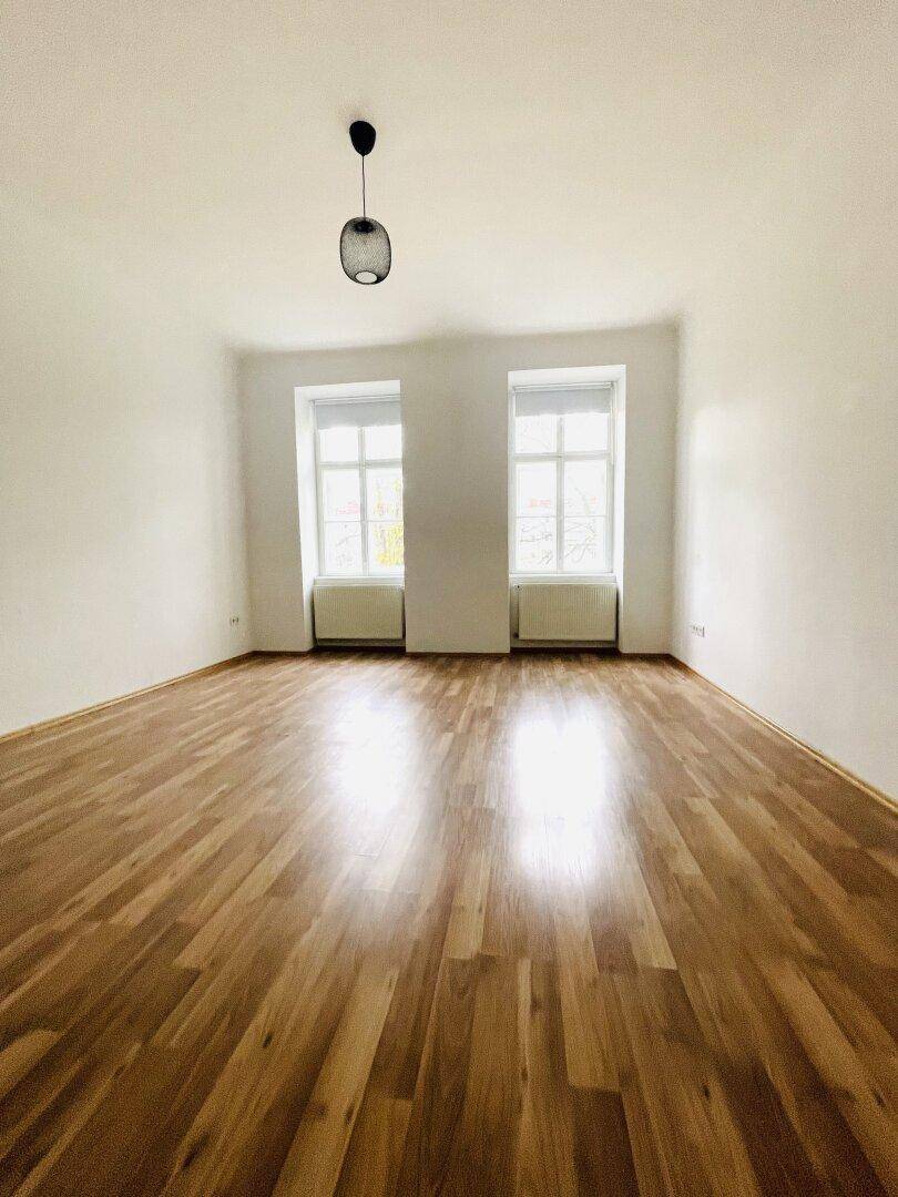 Helles, leeres Zimmer mit glänzendem Parkettboden und zwei Fenstern.