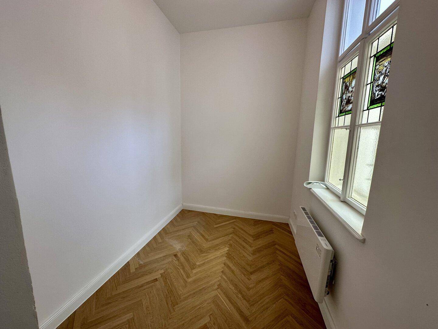 Kleines, helles Zimmer mit Fischgrätparkett und einem Fenster mit Buntglasdetails.