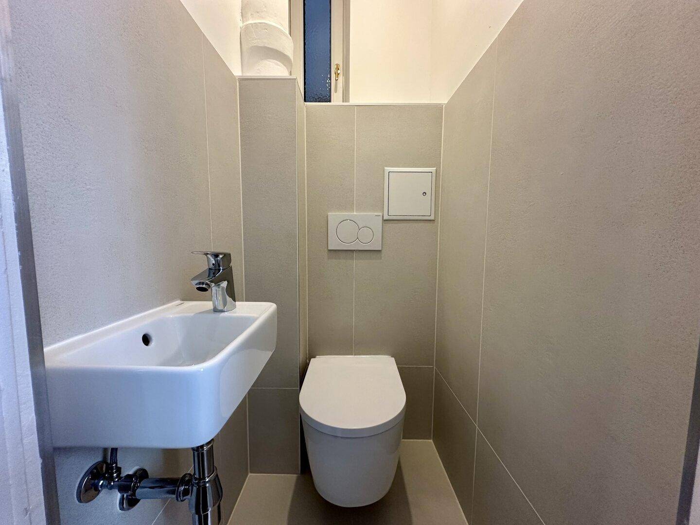 Funktionale Gästetoilette mit Wand-WC, kleinem Waschbecken und modernen Fliesen.