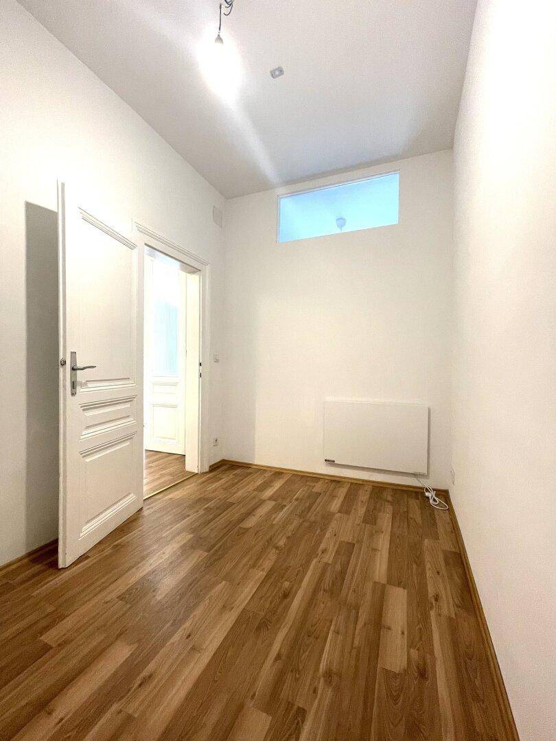 Kleiner Raum mit Holzboden, weißen Wänden und einem hohen Fenster, ideal als Schlaf- oder Arbeitszimmer.