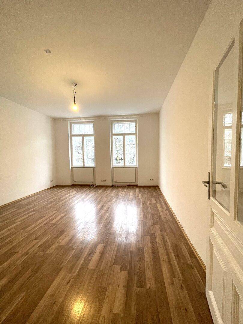 Geräumiges Zimmer mit glänzendem Parkettboden und viel Tageslicht durch die Fenster.