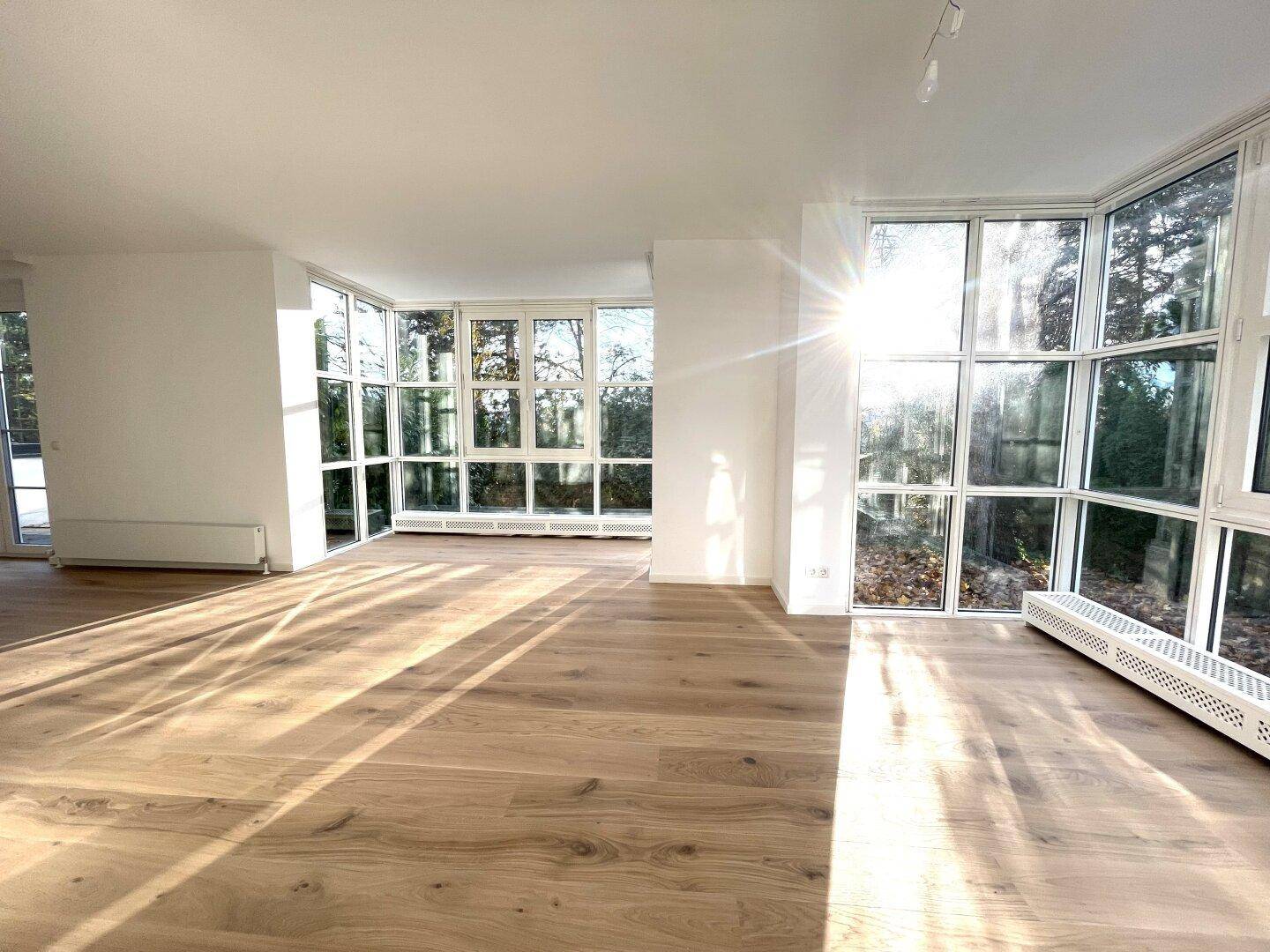 Heller, leerer Wohnbereich mit großen Fenstern, Holzboden und viel natürlichem Sonnenlicht.