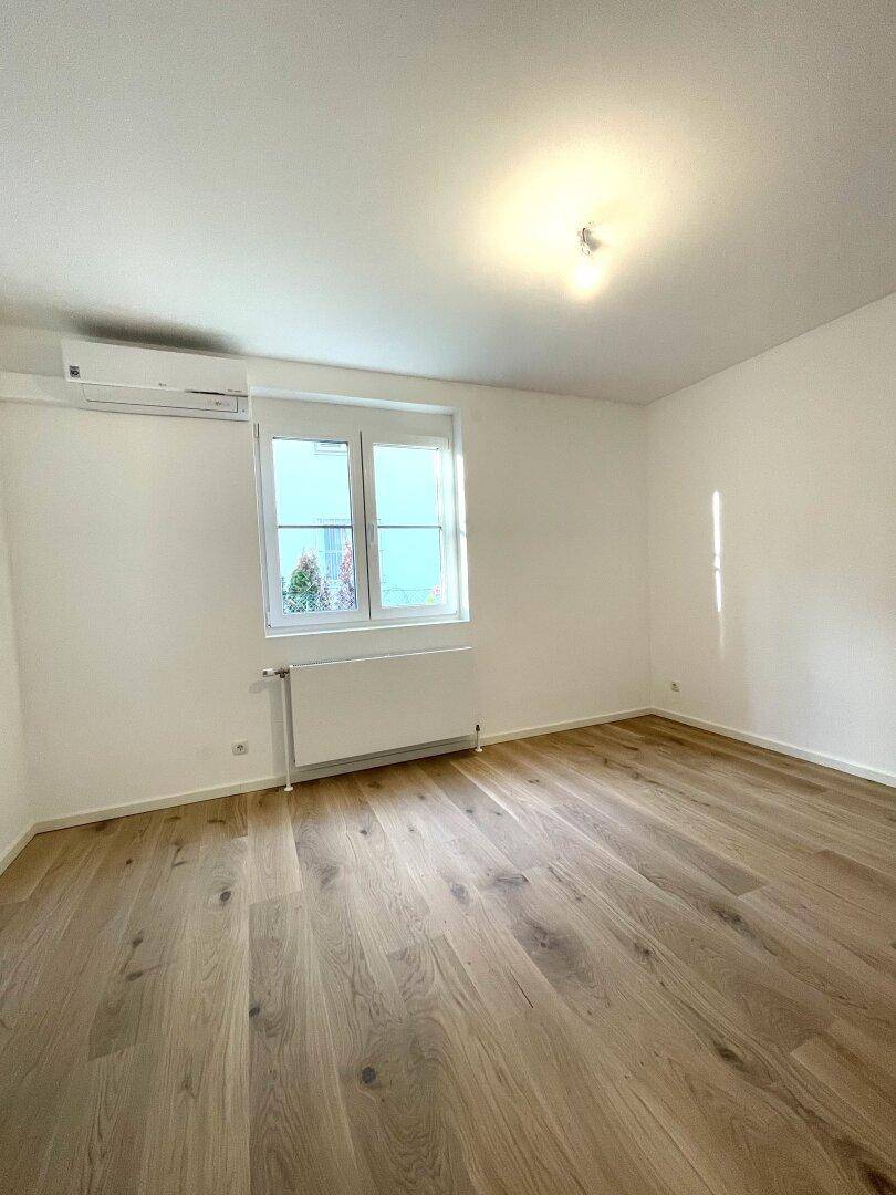 Helles Zimmer mit Holzboden, Fenster und installierter Klimaanlage für angenehmes Raumklima.
