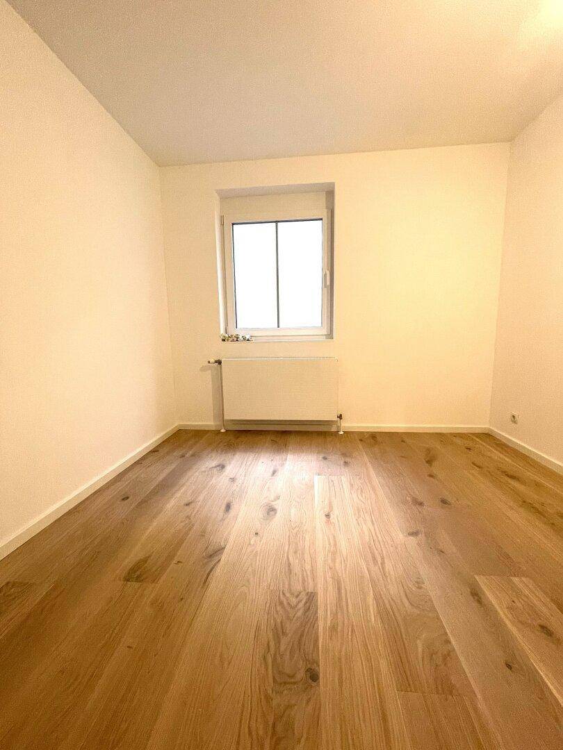 Leeres Zimmer mit hellem Holzparkettboden, Fenster und Heizkörper.