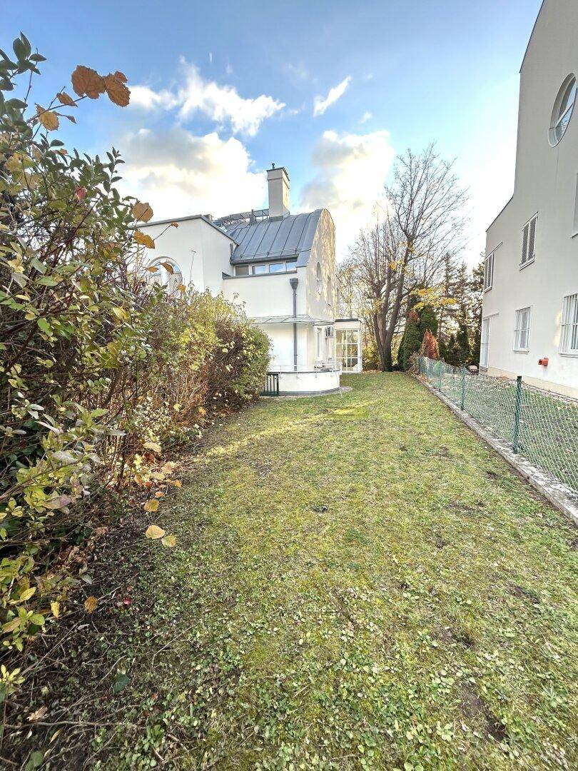 Seitlicher Blick auf das Haus mit gepflegtem Rasen und angrenzenden Büschen unter blauem Himmel.