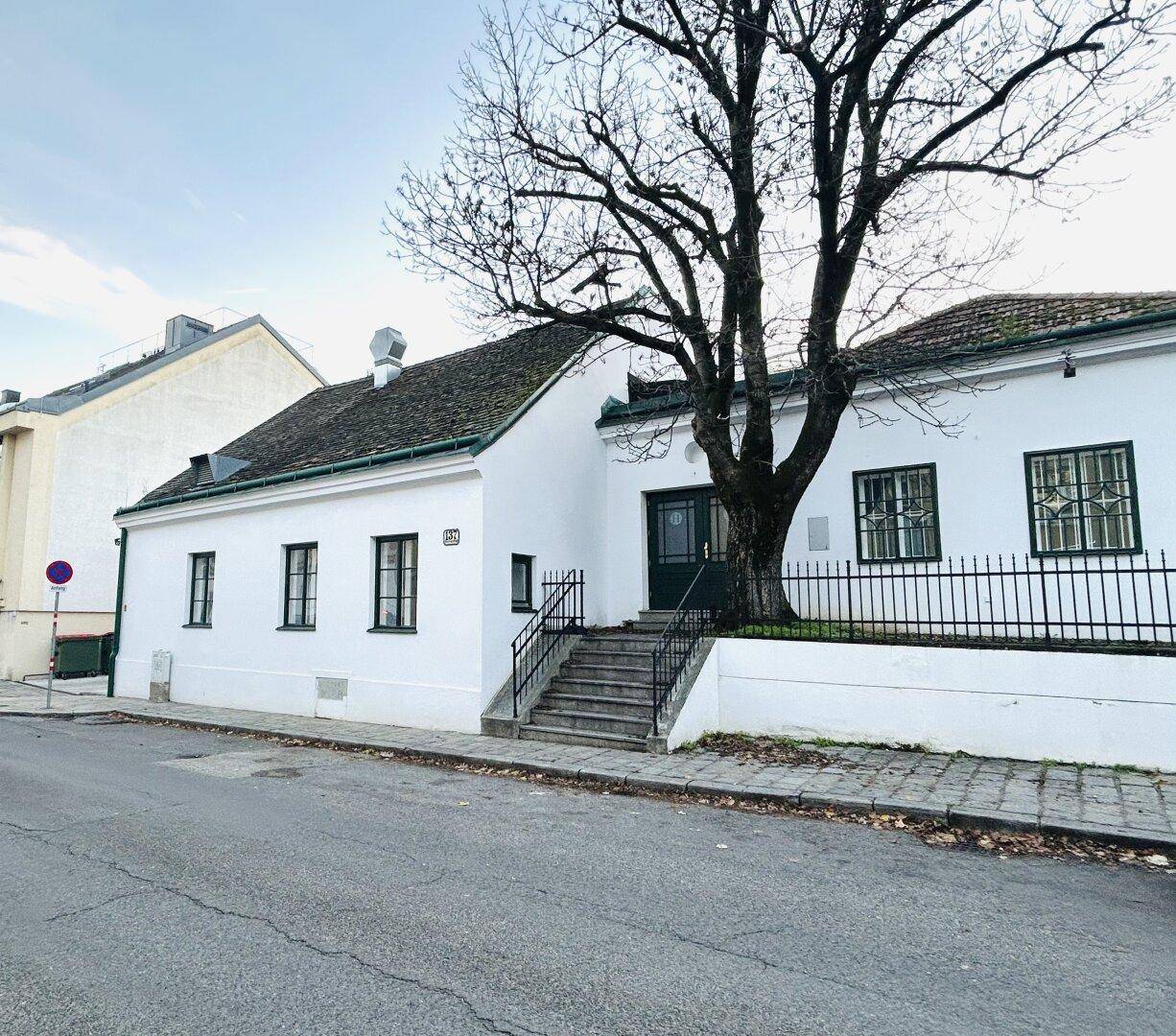 Weißes Gebäude mit dunklem Dach, alter Baum vor dem Eingang und Treppe zur Haustür.