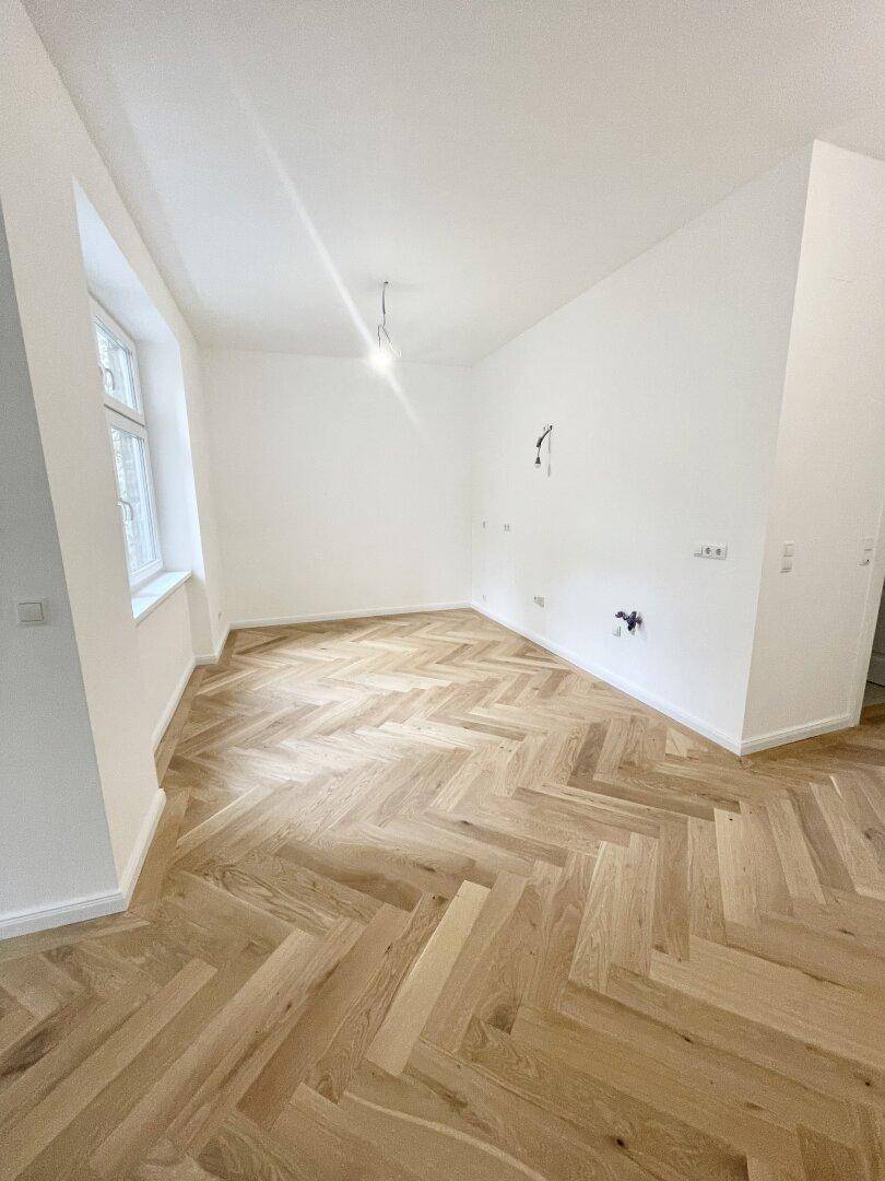 Heller, leerer Raum mit Fischgrätparkett, weißen Wänden und einem Fenster mit Blick nach draußen.