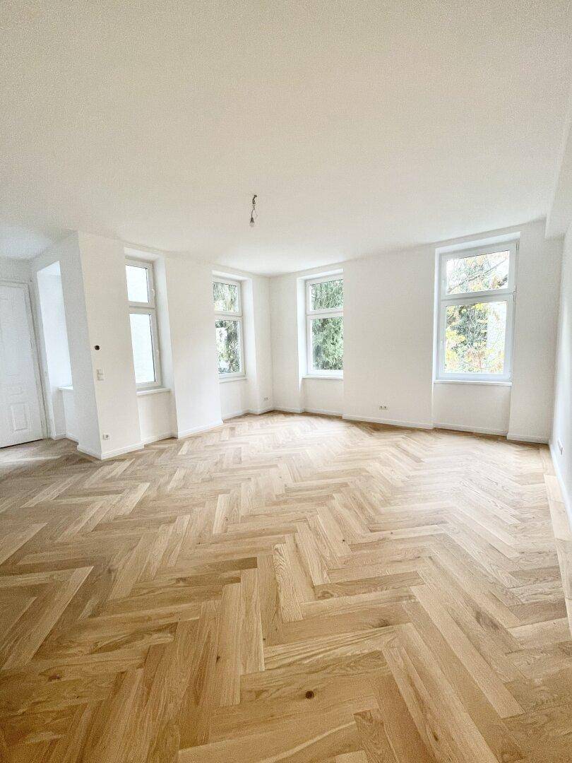 Geräumiger, leerer Raum mit frisch verlegtem Fischgrätparkett, hohen Decken und vielen Fenstern.
