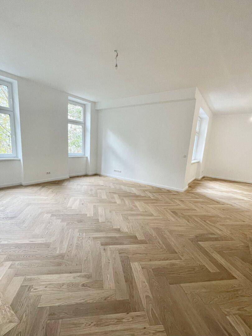 Leerer Raum mit Fischgrätparkett, weißen Wänden und mehreren Fenstern, die viel Licht hereinlassen.