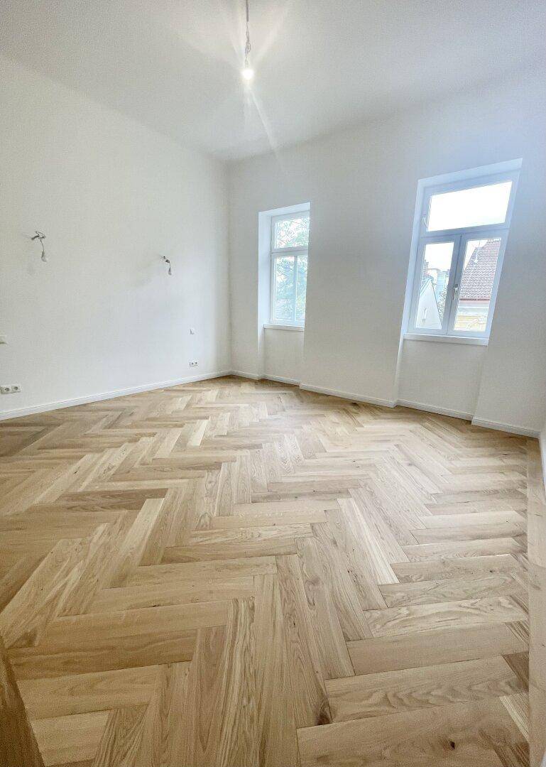 Heller, leerer Raum mit Fischgrätparkett, weißen Wänden und einem Fenster mit Blick nach draußen.