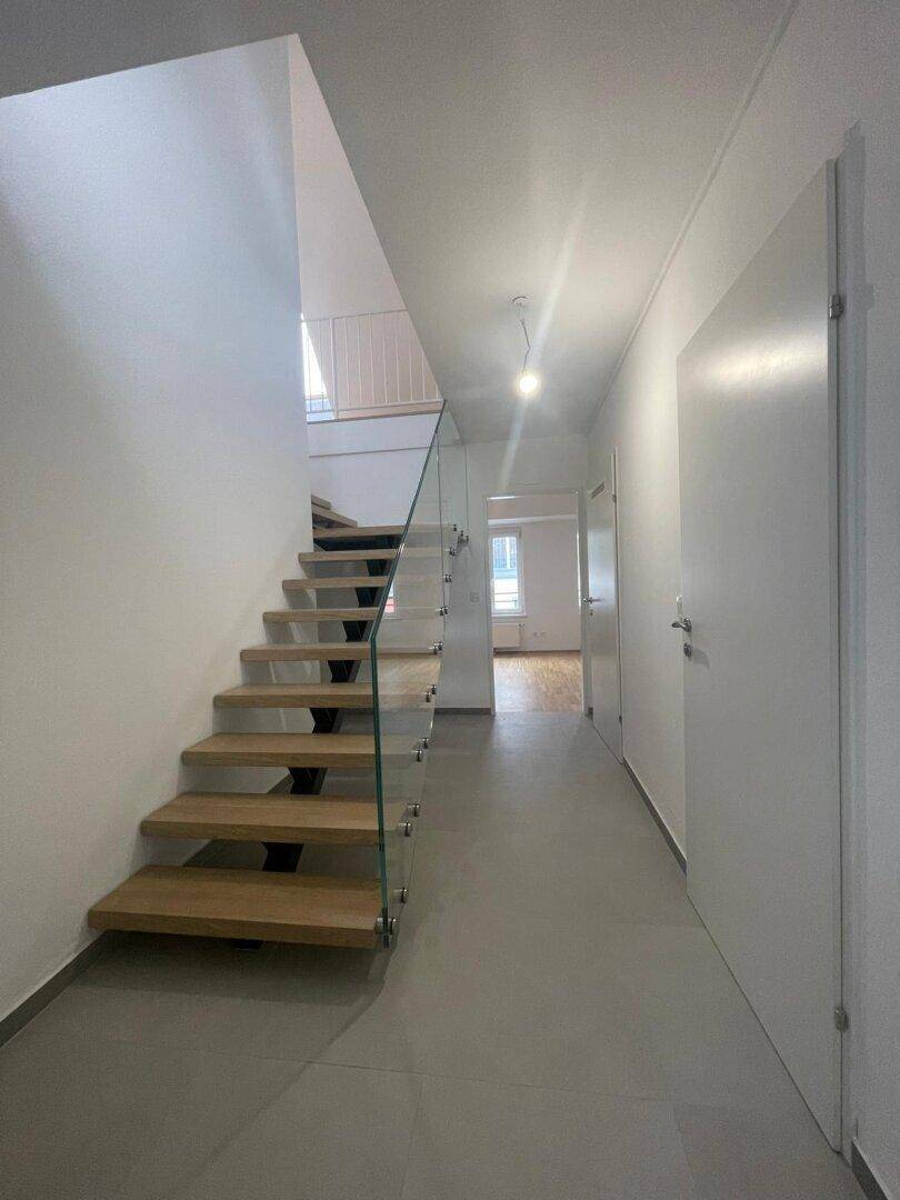 Moderner Flur mit Holztreppe, Glasgeländer und hellem Fliesenboden.
