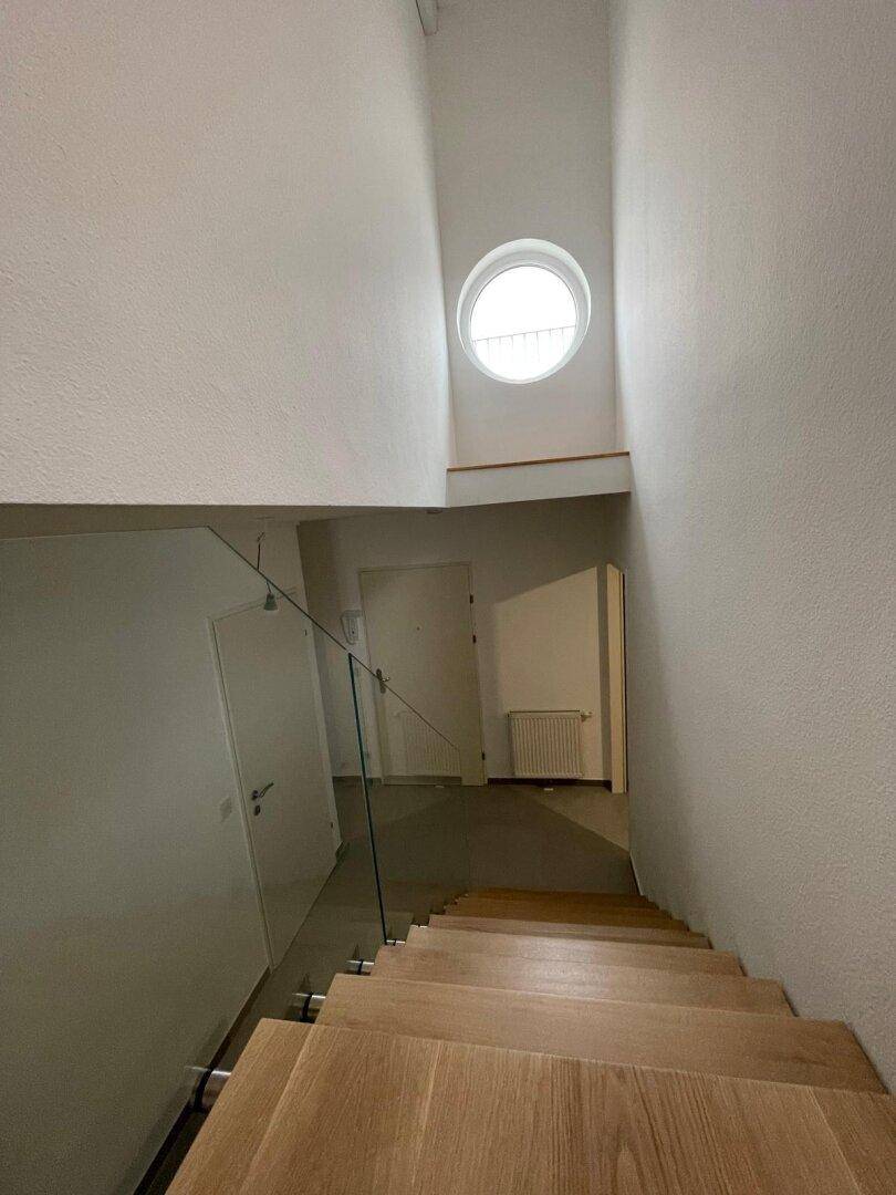 Blick von oben auf die moderne Holztreppe mit Glasgeländer und einem runden Fenster.