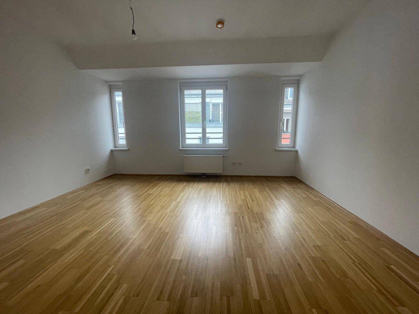 Geräumiges Zimmer mit Holzboden und drei Fenstern, die viel Licht hereinlassen.