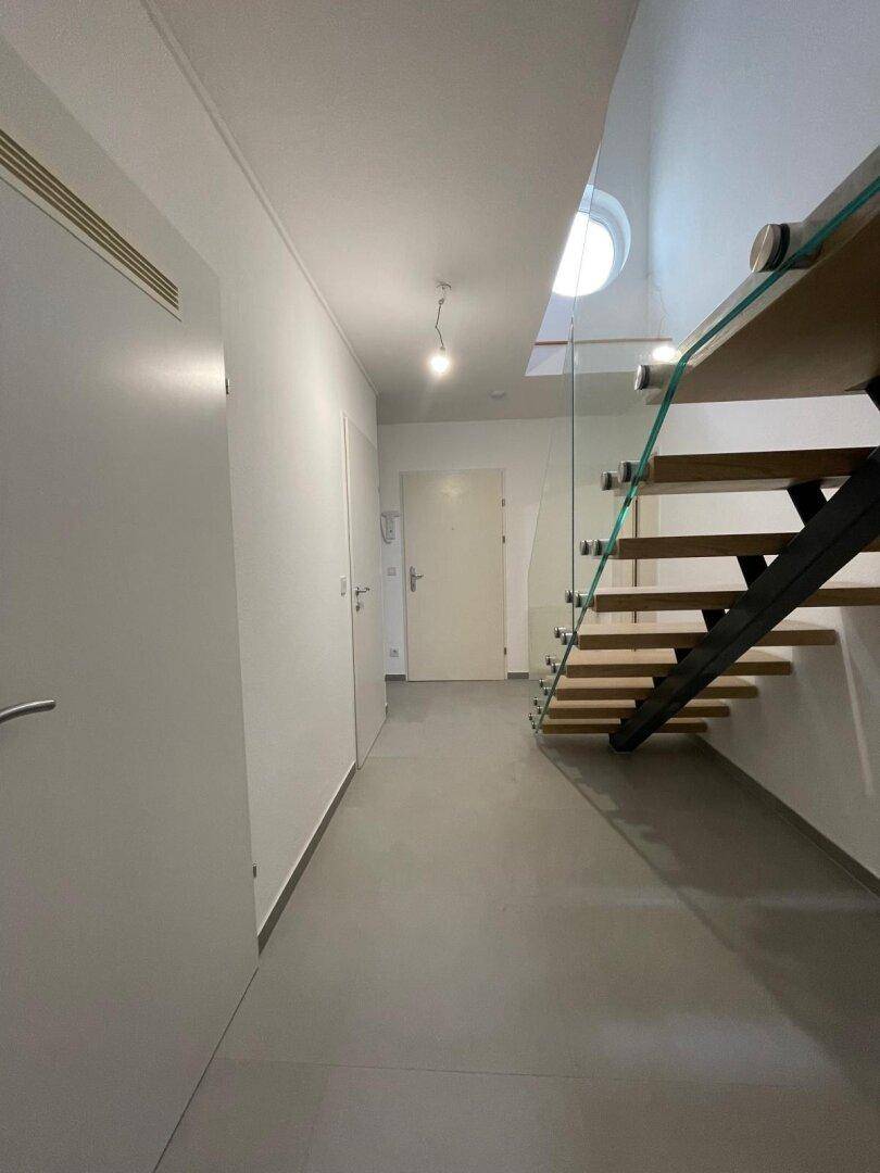 Heller Flur mit grauem Fliesenboden und moderner Holztreppe mit Glasgeländer.